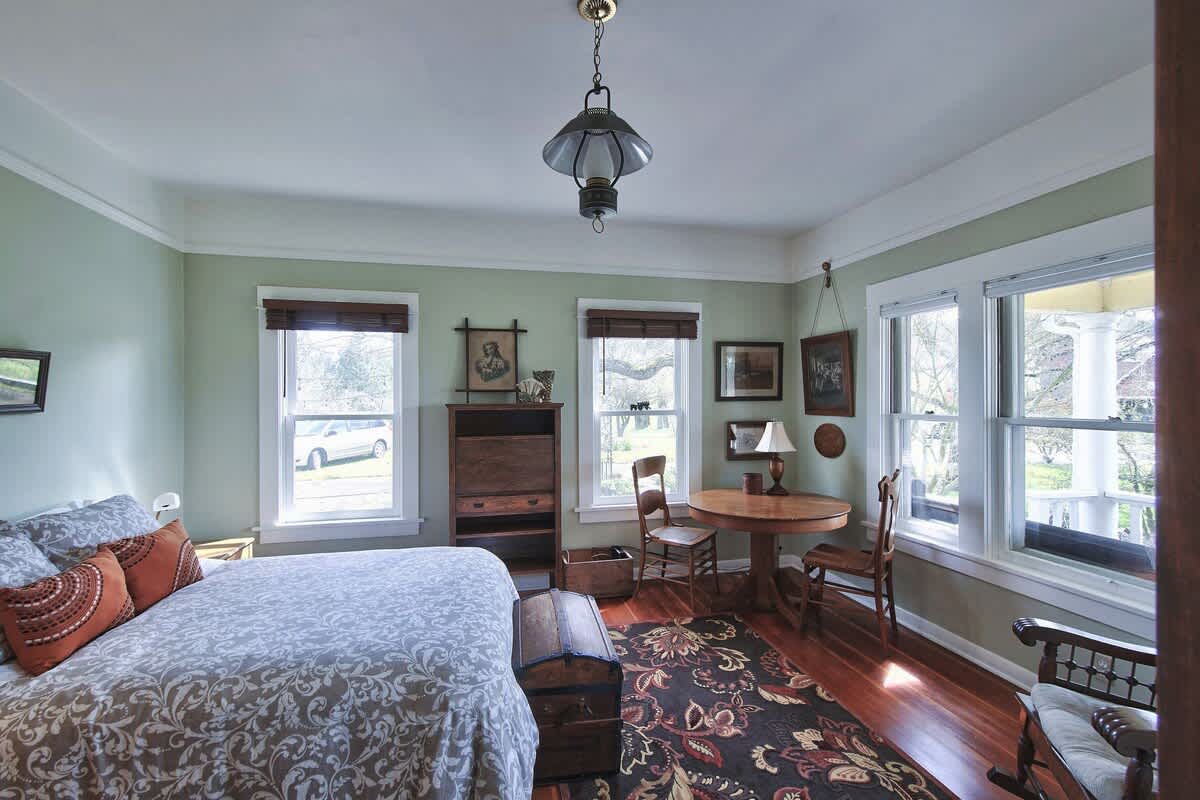 A cozy bedroom with light green walls, hardwood floors, a bed with a patterned duvet, and a small round wooden table with two chairs. Multiple windows offer views of the outdoors.