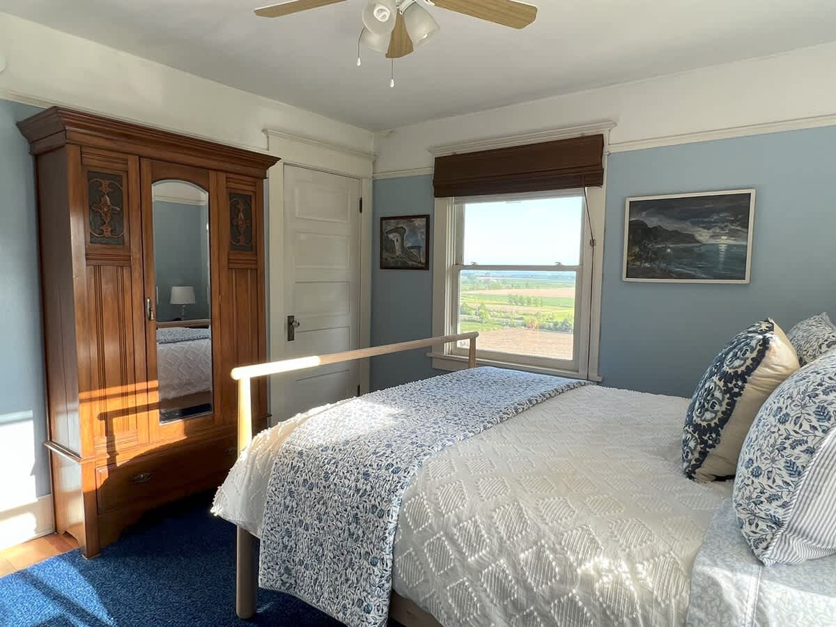 Another view of the blue-walled bedroom, showcasing a large wooden armoire with a mirror, the light wood bed frame with blue and white bedding, and a window with a view of green fields. Sunlight streams into the room.