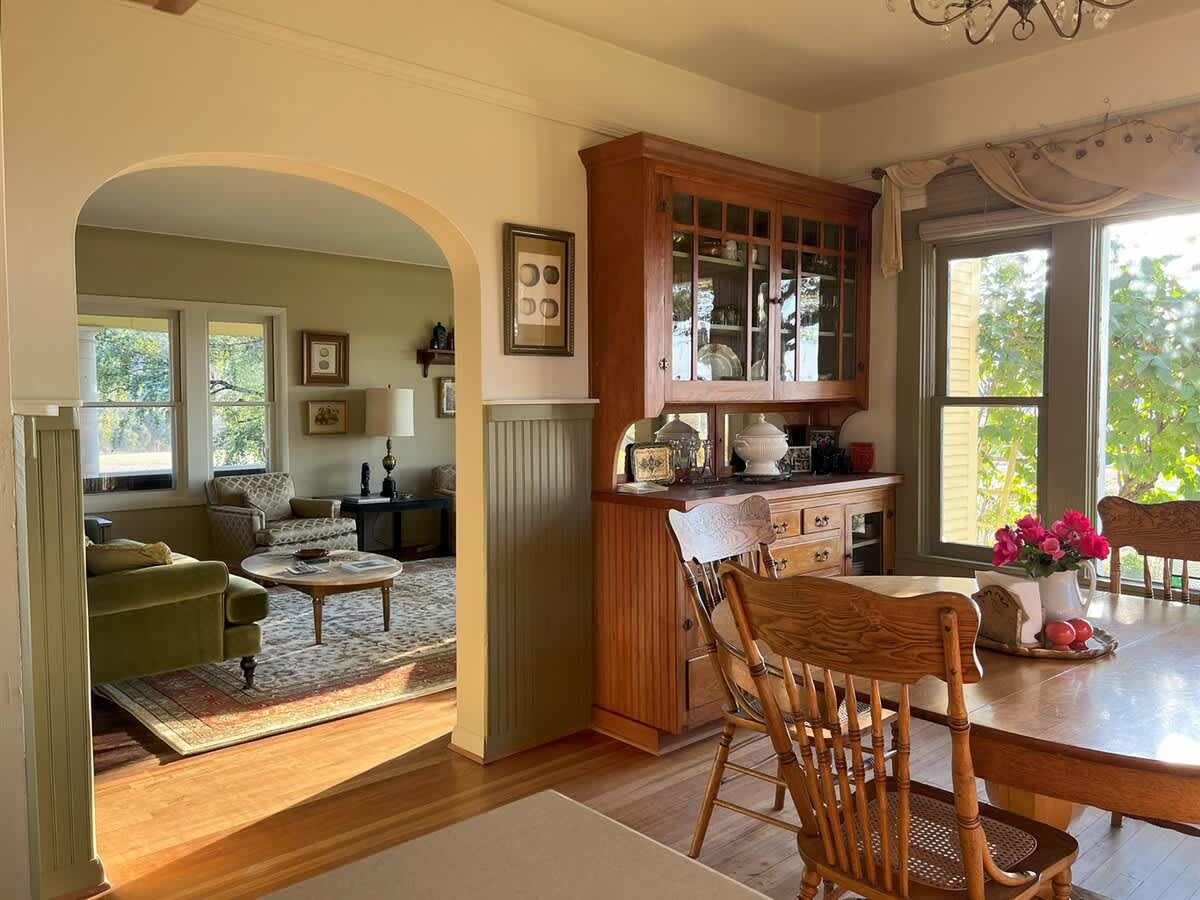 Bright dining room with a round wooden table and matching chairs, a large wooden hutch, and an arched doorway providing a glimpse into the adjacent living room, all set on polished hardwood floors.