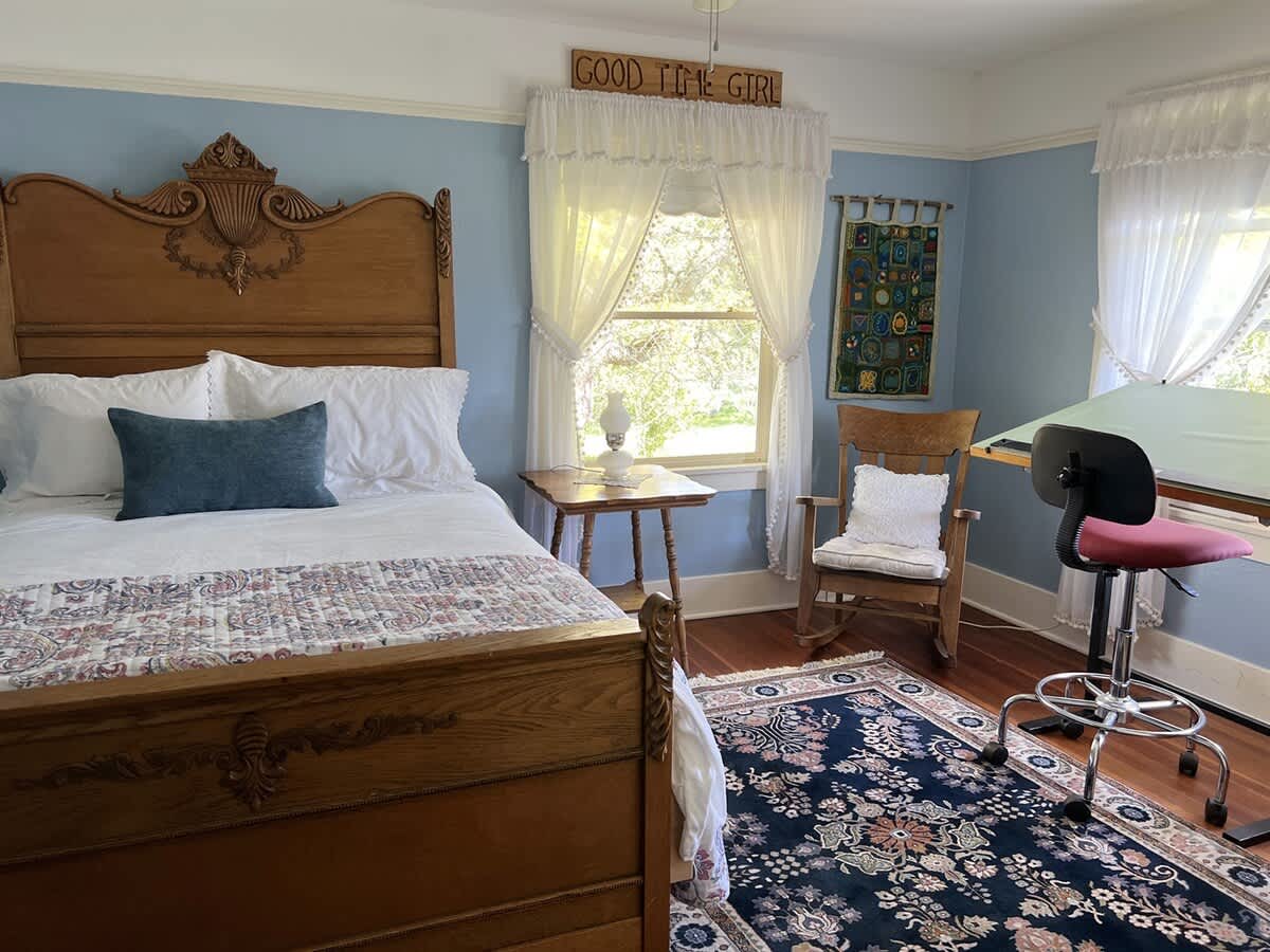 A bright bedroom with light blue walls, a large wooden bed frame, a rocking chair, and a drafting table by a window with sheer white curtains.
