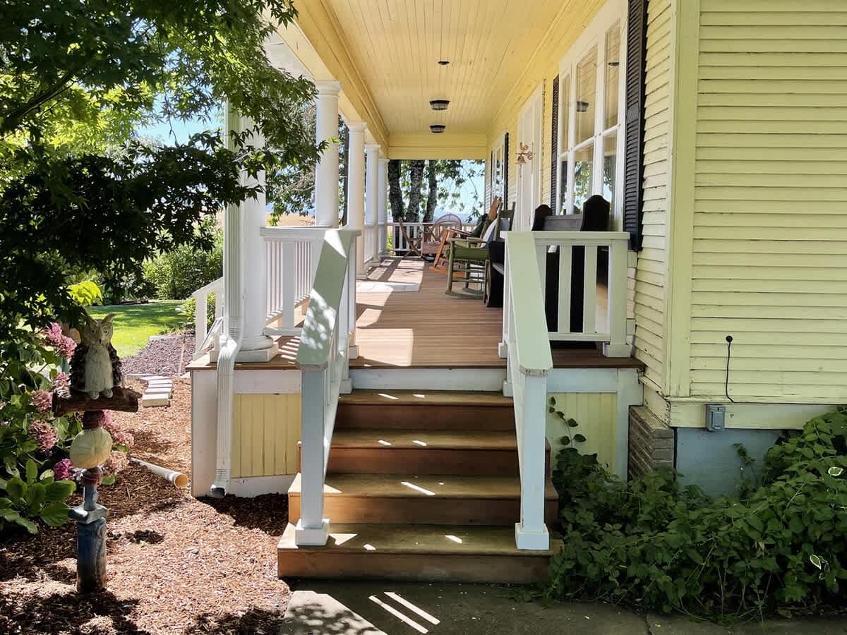 A long wrap-around porch with classic white columns and railings, featuring wooden steps and various seating arrangements, surrounded by lush green landscaping and shaded by trees.