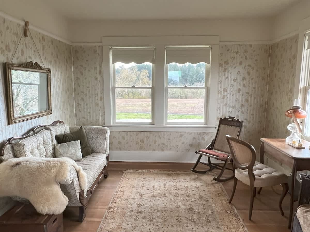 A cozy living room with patterned wallpaper, light wood floors, a vintage-style sofa, a rocking chair, and a wooden desk. Large windows overlook the outdoor scenery.