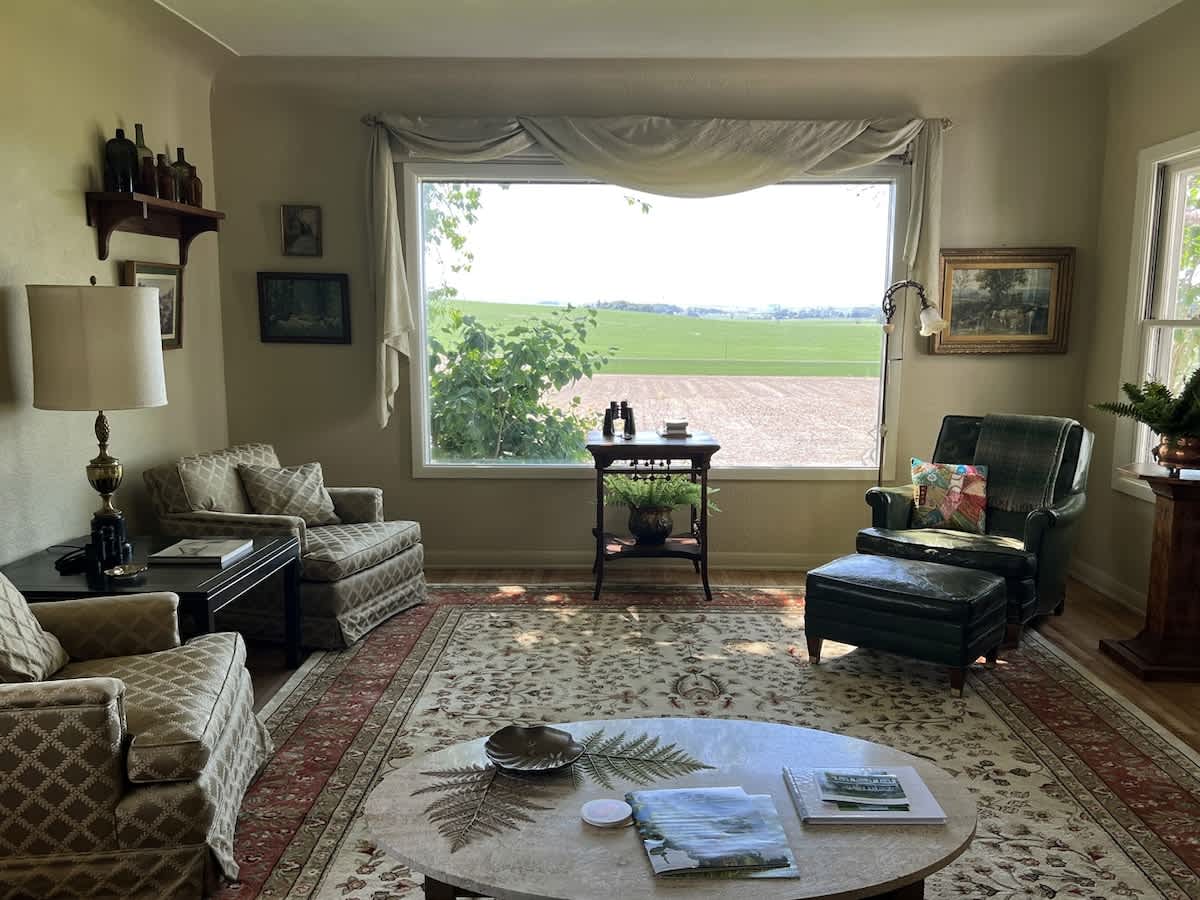 Cozy living room featuring a patterned area rug, a green armchair with an ottoman, and a beige sofa, all centered around a large window offering expansive views of rural fields.
