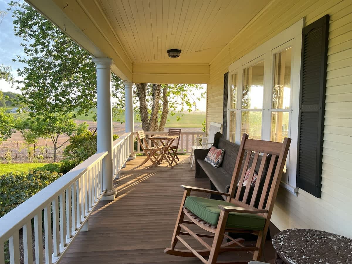 A spacious wooden front porch with white railings, featuring a wooden rocking chair with a green cushion, a dark wooden bench, and a small round table with chairs, overlooking a green landscape.