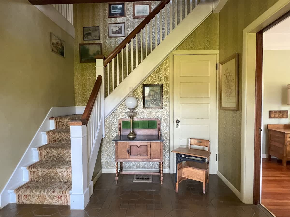 A classic entryway with patterned wallpaper and tiled floors, featuring a carpeted staircase with white banisters, a vintage wooden console table, and an antique school desk.