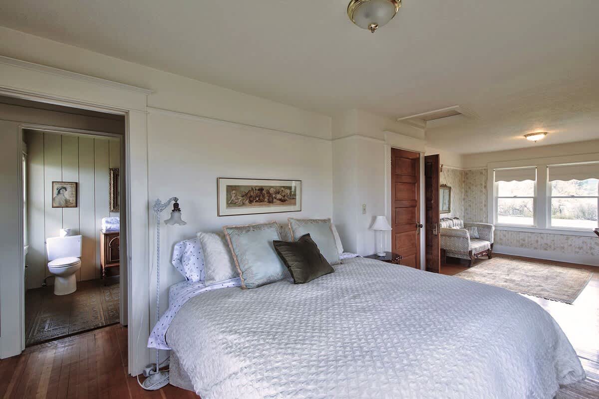 A bright bedroom with a large white quilted bed, light blue decorative pillows, and a view into an adjacent bathroom with a toilet and vanity.