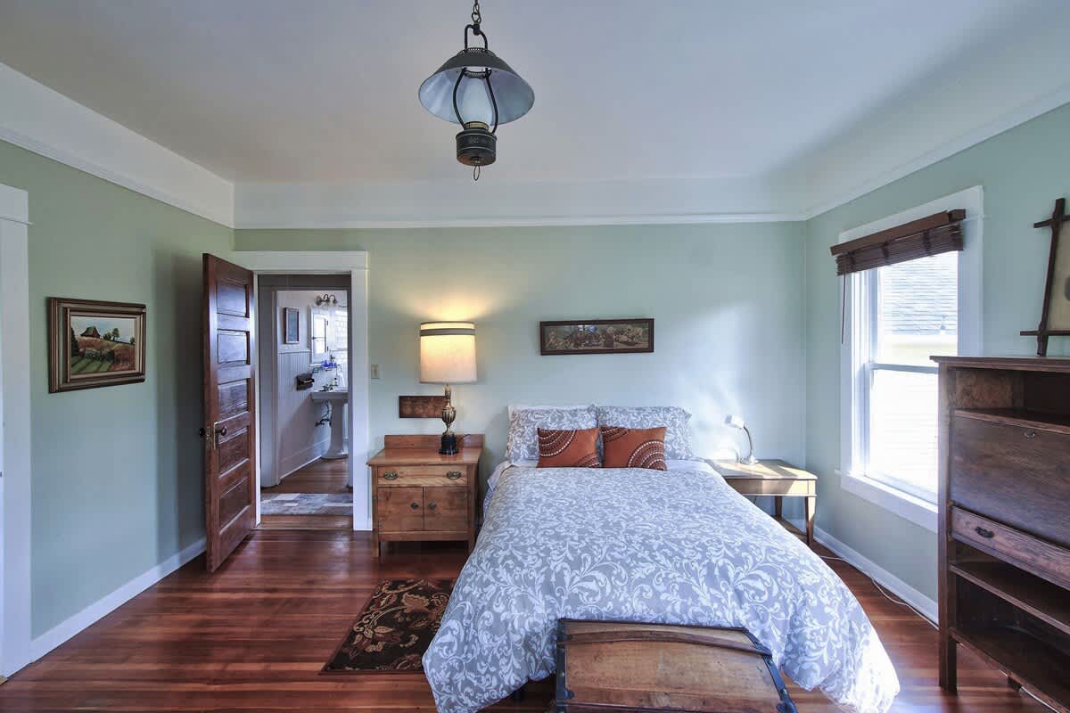 View of a bedroom with light green walls and polished hardwood floors, featuring a bed with a patterned duvet, a wooden nightstand with a lamp, and an open doorway leading to a bathroom.