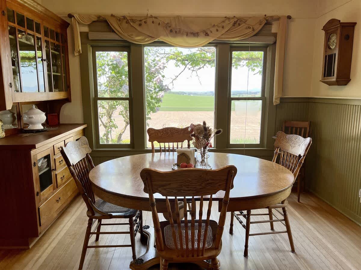 Charming dining room centered around a round wooden table with classic chairs, positioned by a wide window offering picturesque views of the expansive rural landscape and distant fields.