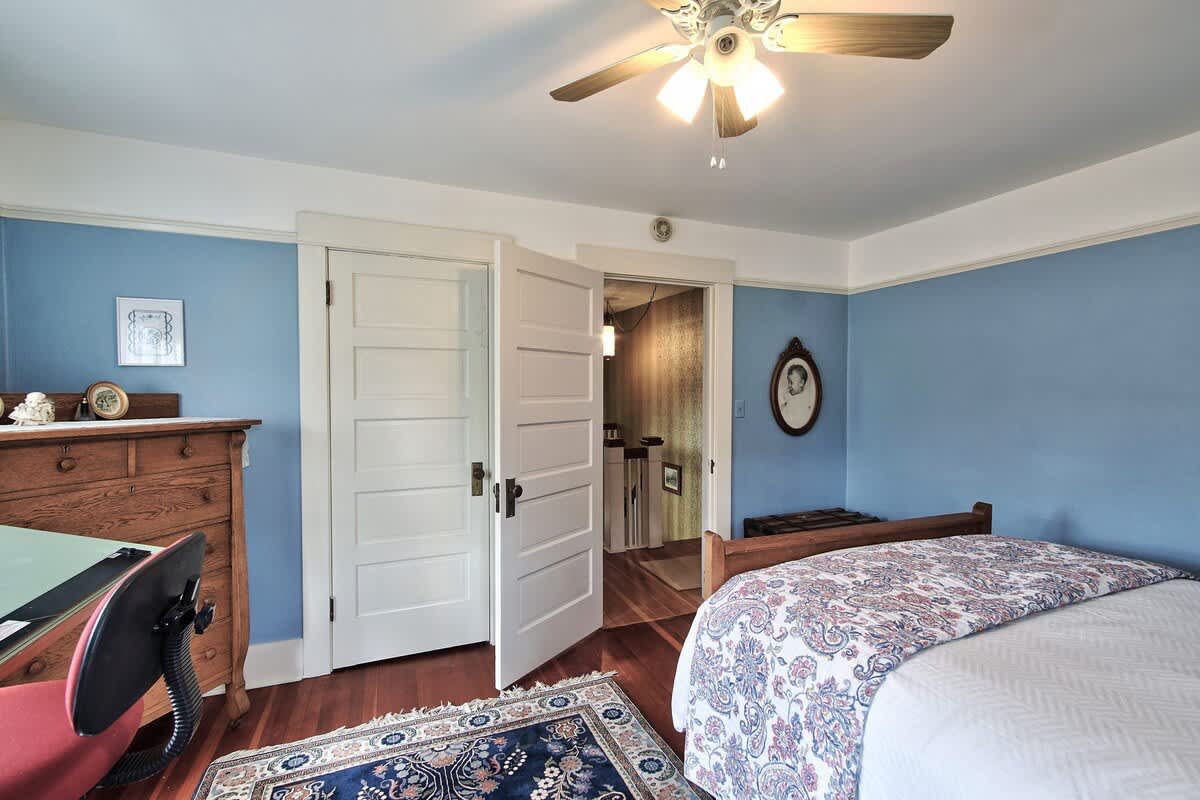 A cozy bedroom with light blue walls, a wooden bed, a drafting table, and an open doorway leading to a hallway with patterned wallpaper.