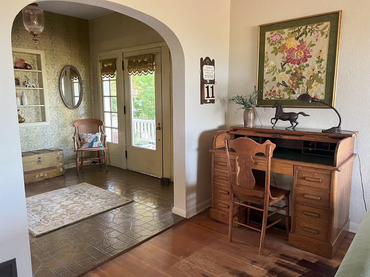 A cozy office nook with hardwood floors, featuring a vintage roll-top desk with a wooden chair, a decorative lamp, and a framed floral tapestry on the wall, adjacent to the foyer.