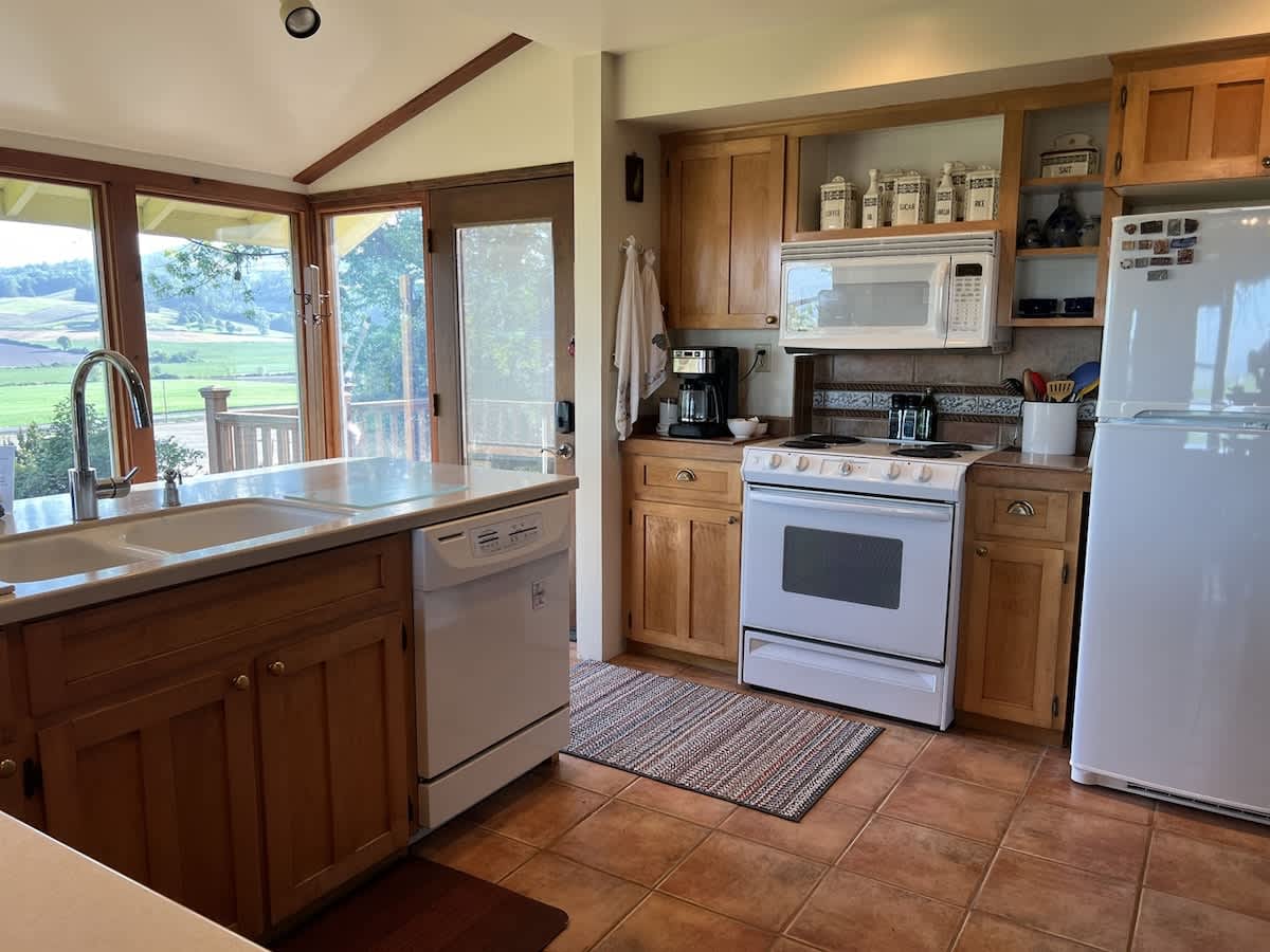 A bright kitchen with light wood cabinets, white appliances including a stove, microwave, and refrigerator, and a large window overlooking a green landscape. The floor is tiled in terracotta.