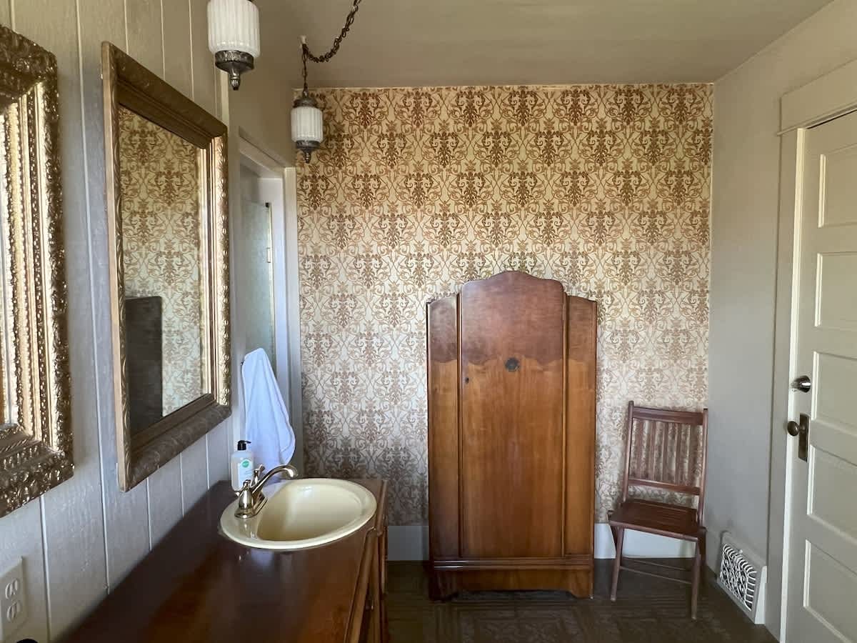 A vintage bathroom featuring a dark wood vanity with a cream sink, ornate mirrors, and a tall wooden wardrobe, complemented by patterned wallpaper and floor tiles.