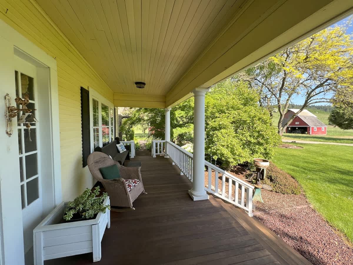 The inviting front porch features yellow siding, white columns, and a dark wooden floor, with a glimpse of a red barn in the distance, surrounded by vibrant green foliage.