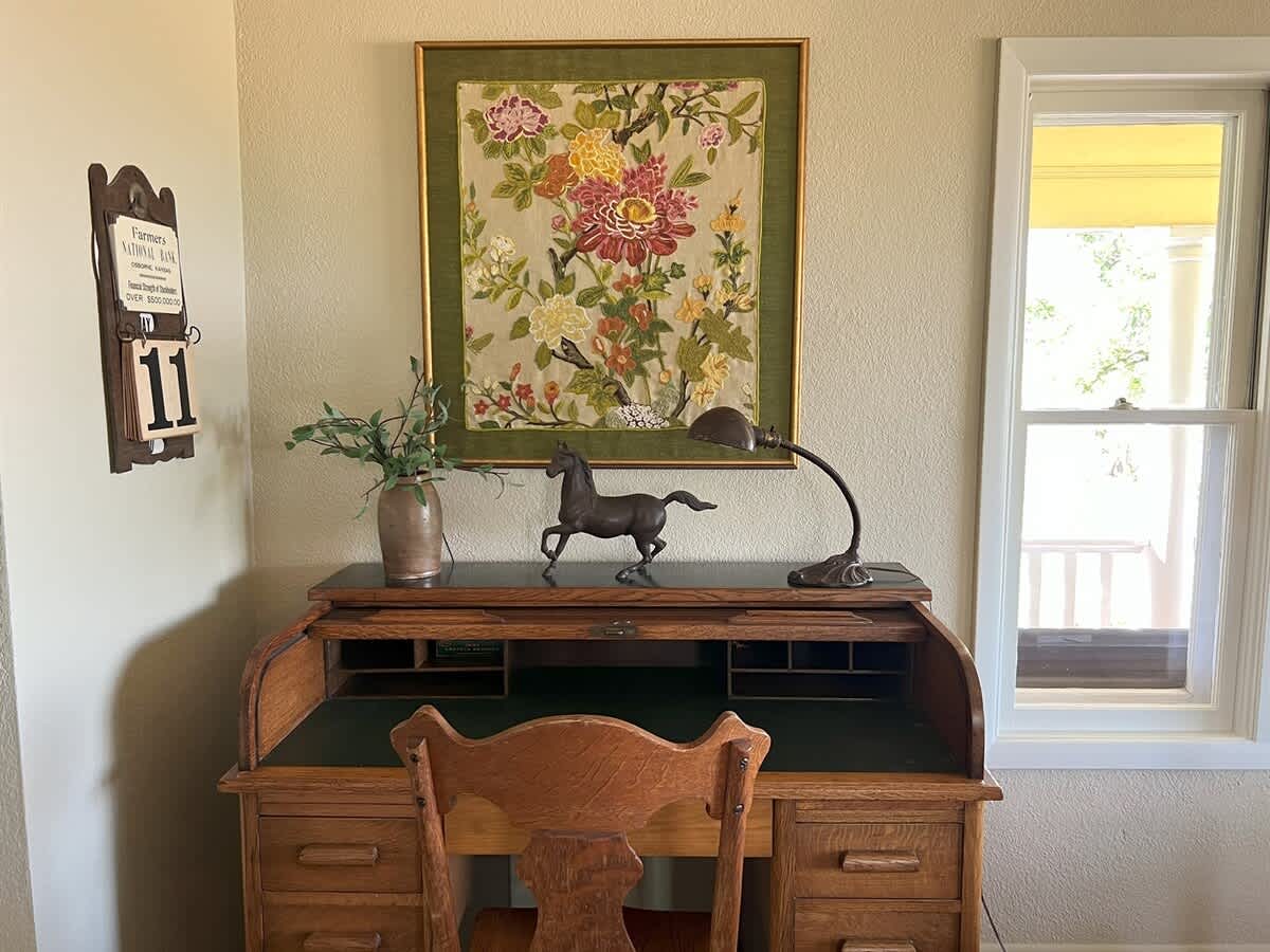 A close-up of a vintage wooden roll-top desk with a green felt surface, adorned with a bronze horse statue and a desk lamp, beneath a framed floral tapestry.