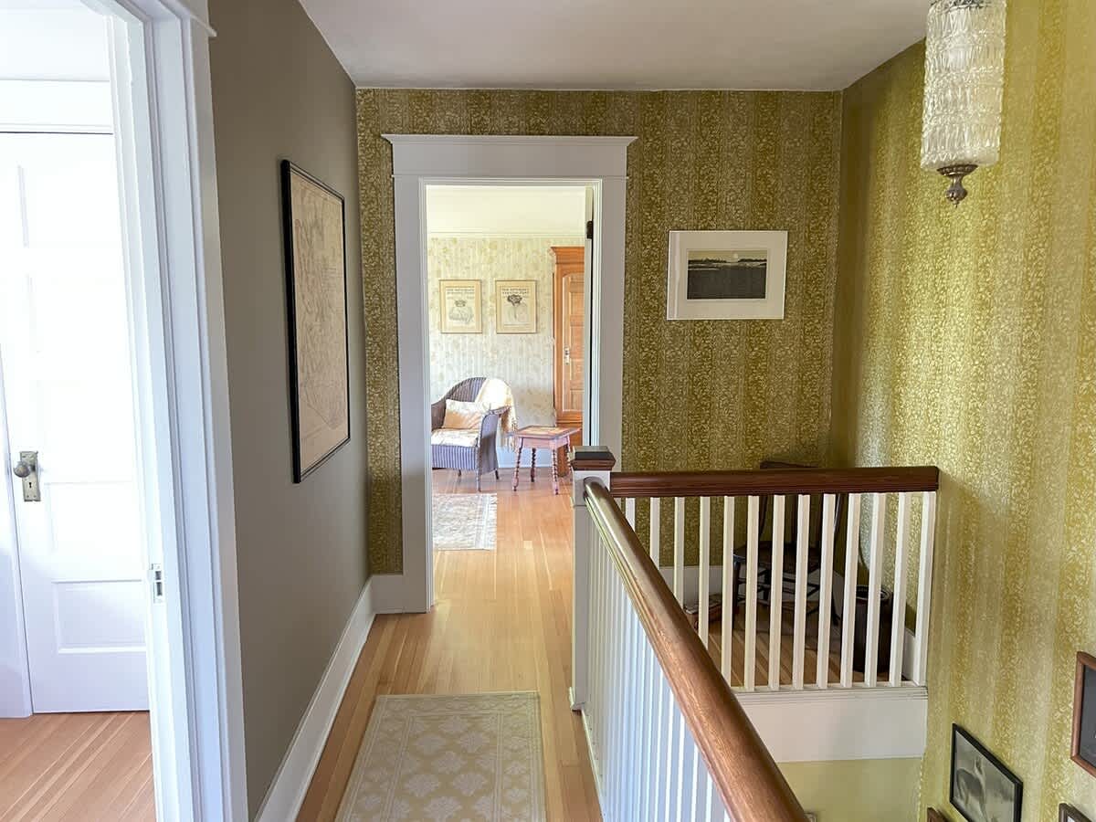 A hallway with light wood floors and patterned green wallpaper, featuring a white banister leading down a staircase and an open doorway to another room.