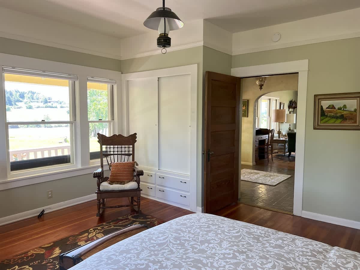 A bright bedroom with light green walls and hardwood floors, featuring a wooden rocking chair, a built-in white closet, and large windows offering a view of the exterior landscape.