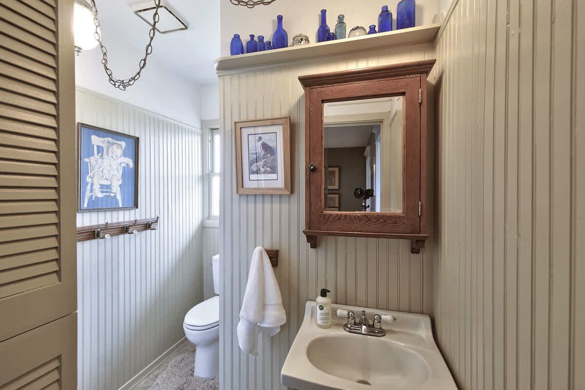 A bright bathroom with light beadboard walls, featuring a white pedestal sink, a toilet, and a wooden medicine cabinet with a mirror. Blue bottles are displayed on a shelf above the mirror.