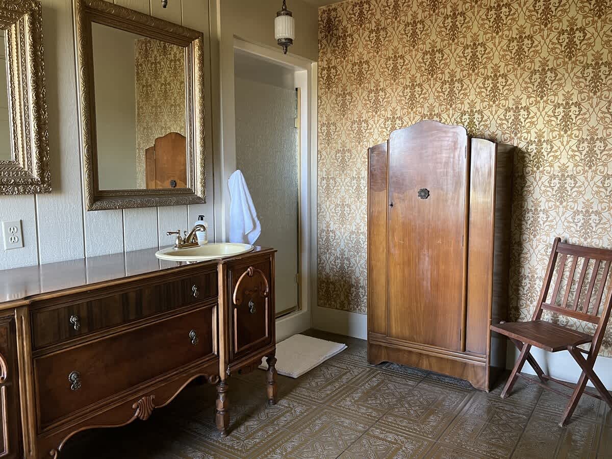 A spacious vintage bathroom with a dark wood vanity, ornate mirrors, a wooden wardrobe, and a visible shower entrance, all set on patterned floor tiles.