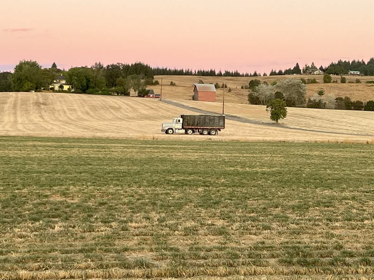 A large white semi-truck with a dark trailer travels along a dirt road through expansive, recently harvested fields, with a red barn and other farm buildings visible on a distant hill under a soft pink and orange sunset sky.