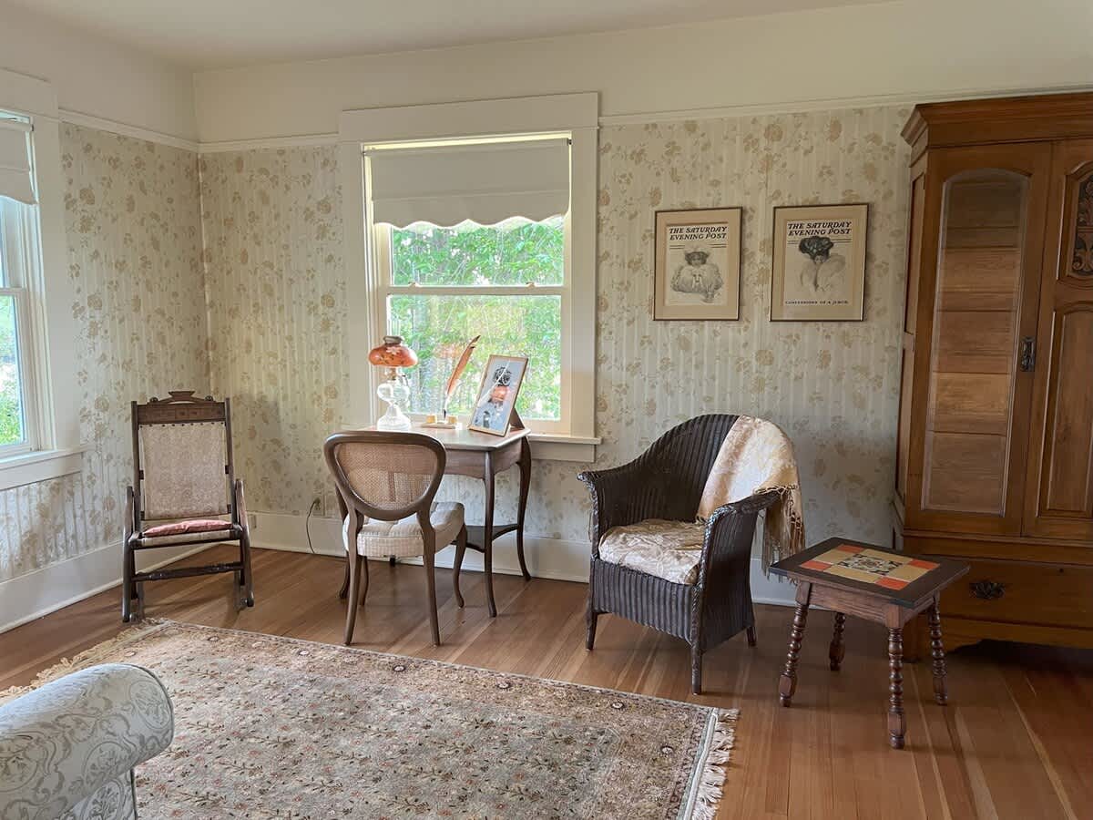 A spacious living room featuring a patterned area rug, a dark wooden armoire, a wicker armchair, and a small writing desk by a window with floral wallpaper.