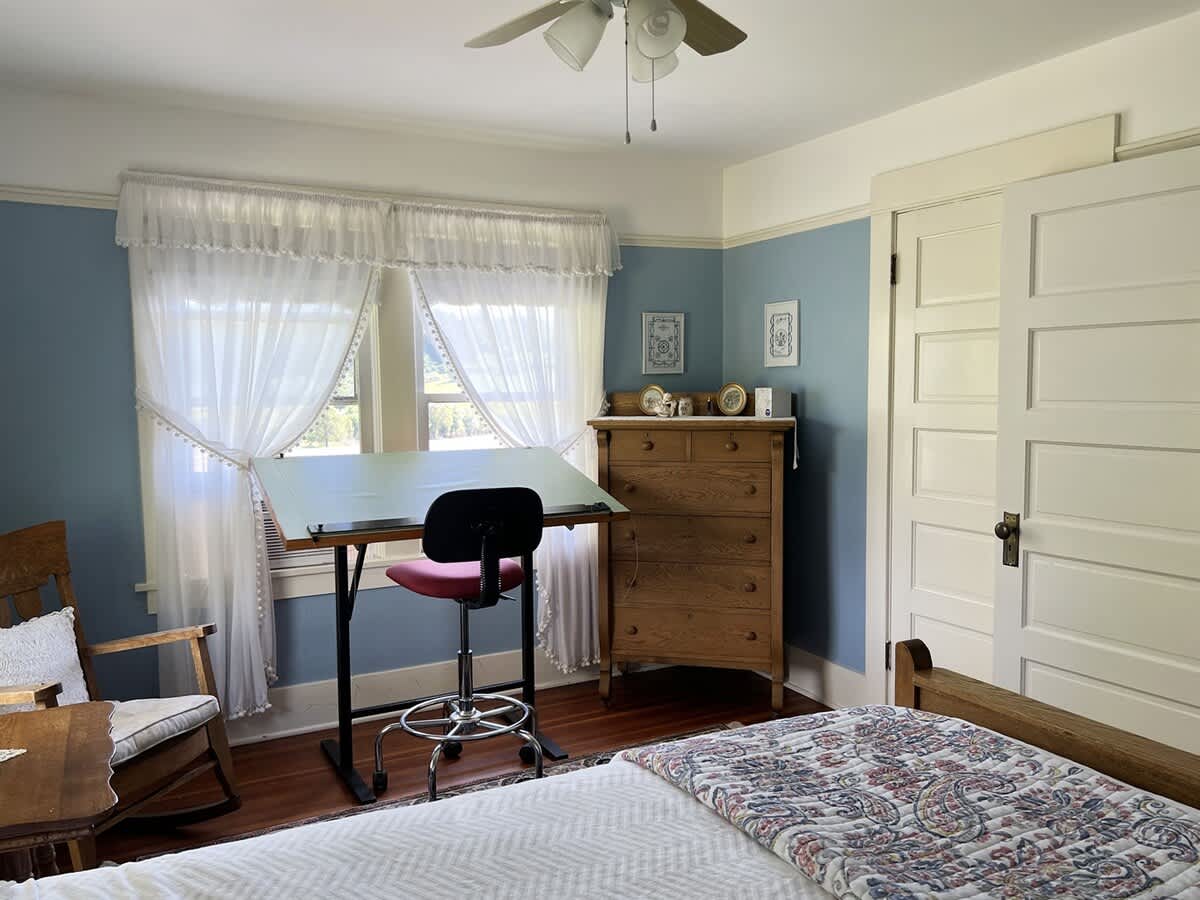 A light-filled bedroom featuring a wooden bed, a drafting table, a wooden dresser, and a window dressed with white sheer curtains, all against light blue walls.
