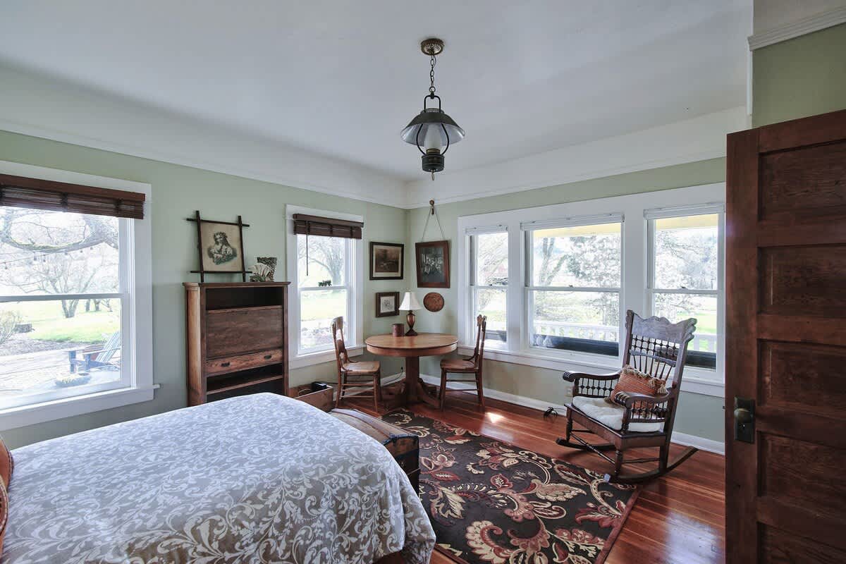 A spacious bedroom with light green walls, hardwood floors, a bed with a patterned duvet, a small round wooden table, and a rocking chair. Multiple windows provide ample natural light.