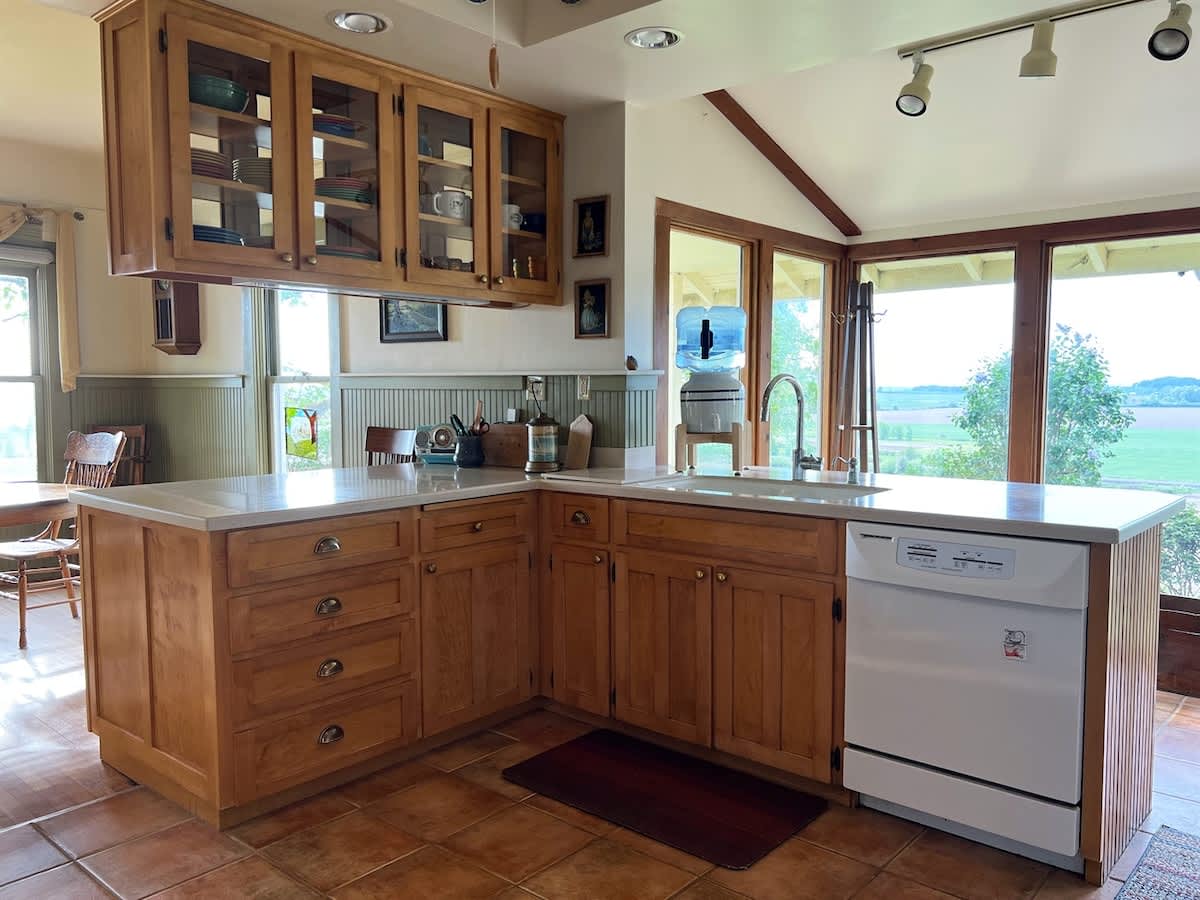Bright kitchen area with light wood cabinets, white countertops, a double sink, and a dishwasher, illuminated by large windows that offer views of the exterior, all set on a practical tiled floor.