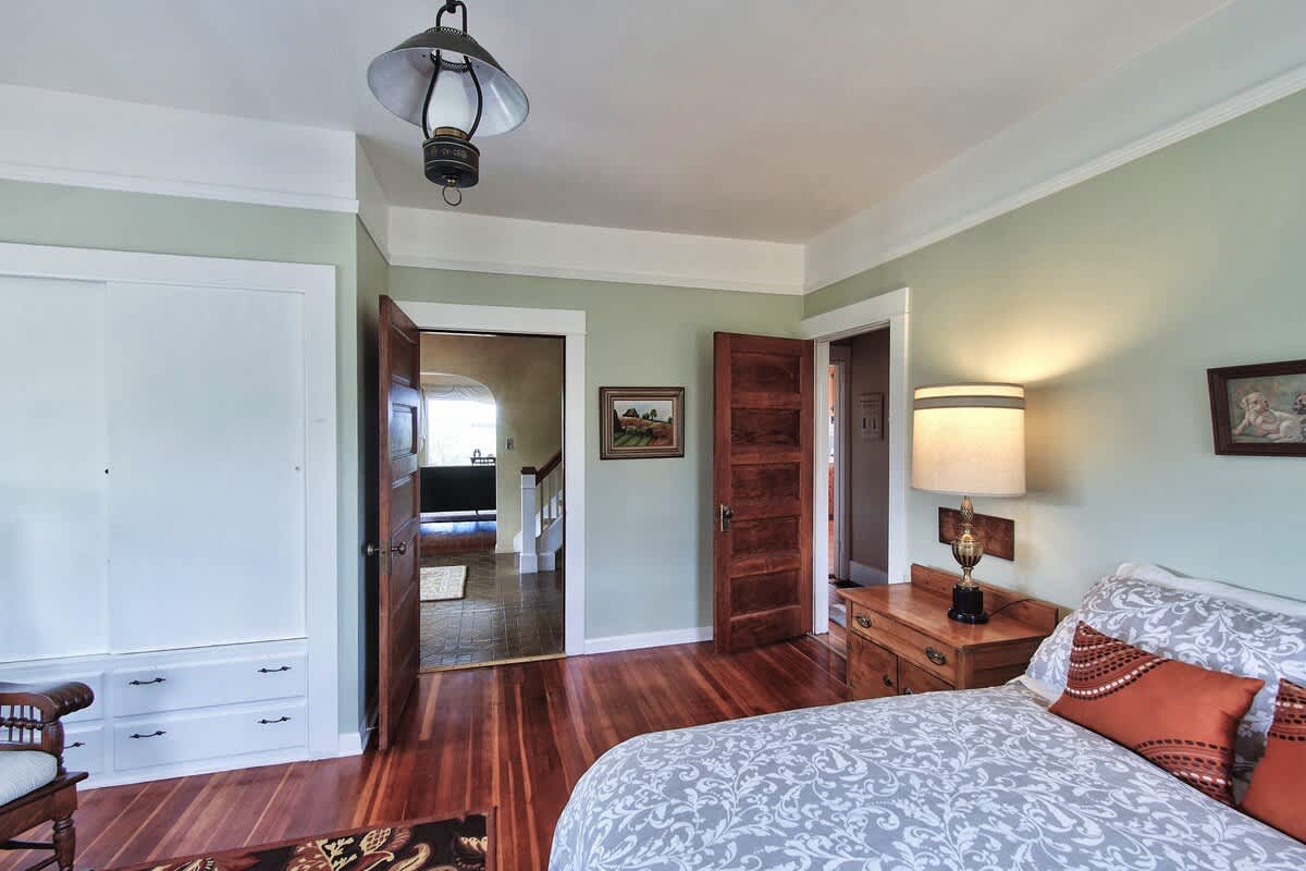 A bedroom with light green walls and hardwood floors, featuring a built-in white closet with sliding doors and drawers, and an open wooden door leading to an adjacent room.