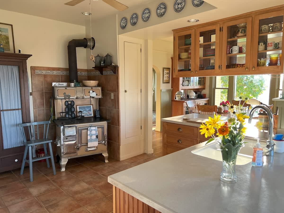 A rustic kitchen area featuring a vintage-style wood-burning stove with a stovepipe, light wood cabinetry, and terracotta tile flooring. A large window provides natural light.
