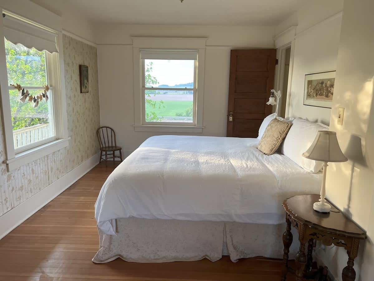 A bright bedroom with white bedding on a large bed, hardwood floors, and two windows offering views of green fields and distant hills. A small wooden chair and a bedside table with a lamp are visible.