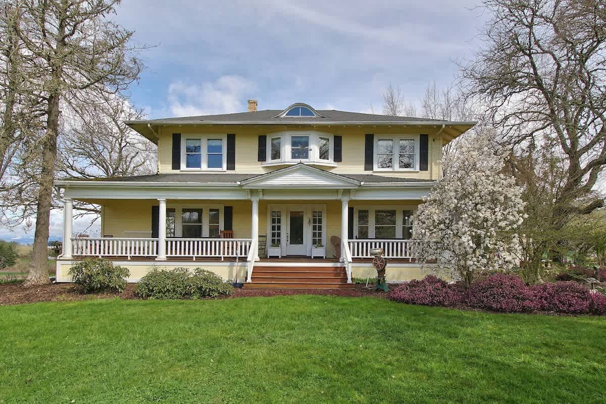 A grand yellow two-story farm home with white trim, black shutters, and a wide wraparound porch with white columns and railings. The house is surrounded by green grass and mature trees, with a bright blue sky overhead.