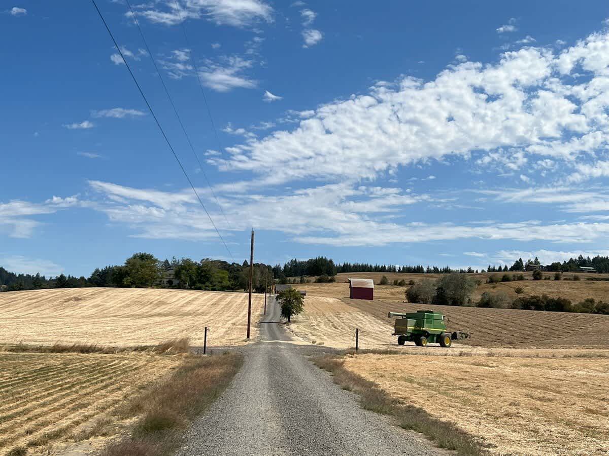 A gravel road ascends through vast, golden harvested fields, with a green combine harvester parked on the right and a red barn visible in the distance, all under a bright blue sky with scattered white clouds.