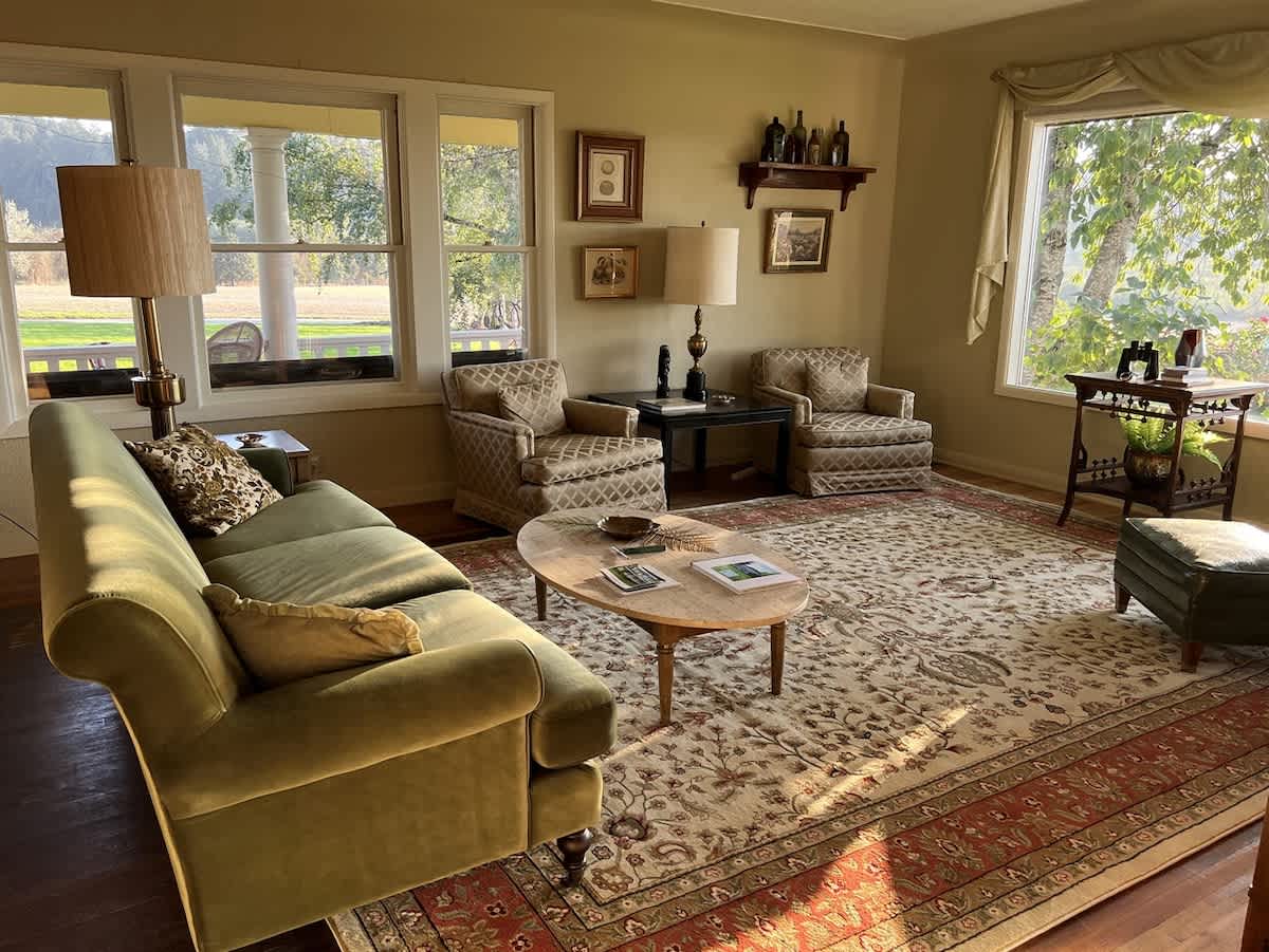 A wide view of a traditional living room with a green velvet sofa, two patterned armchairs, a large ornate rug, and a marble coffee table, illuminated by natural light from multiple windows.