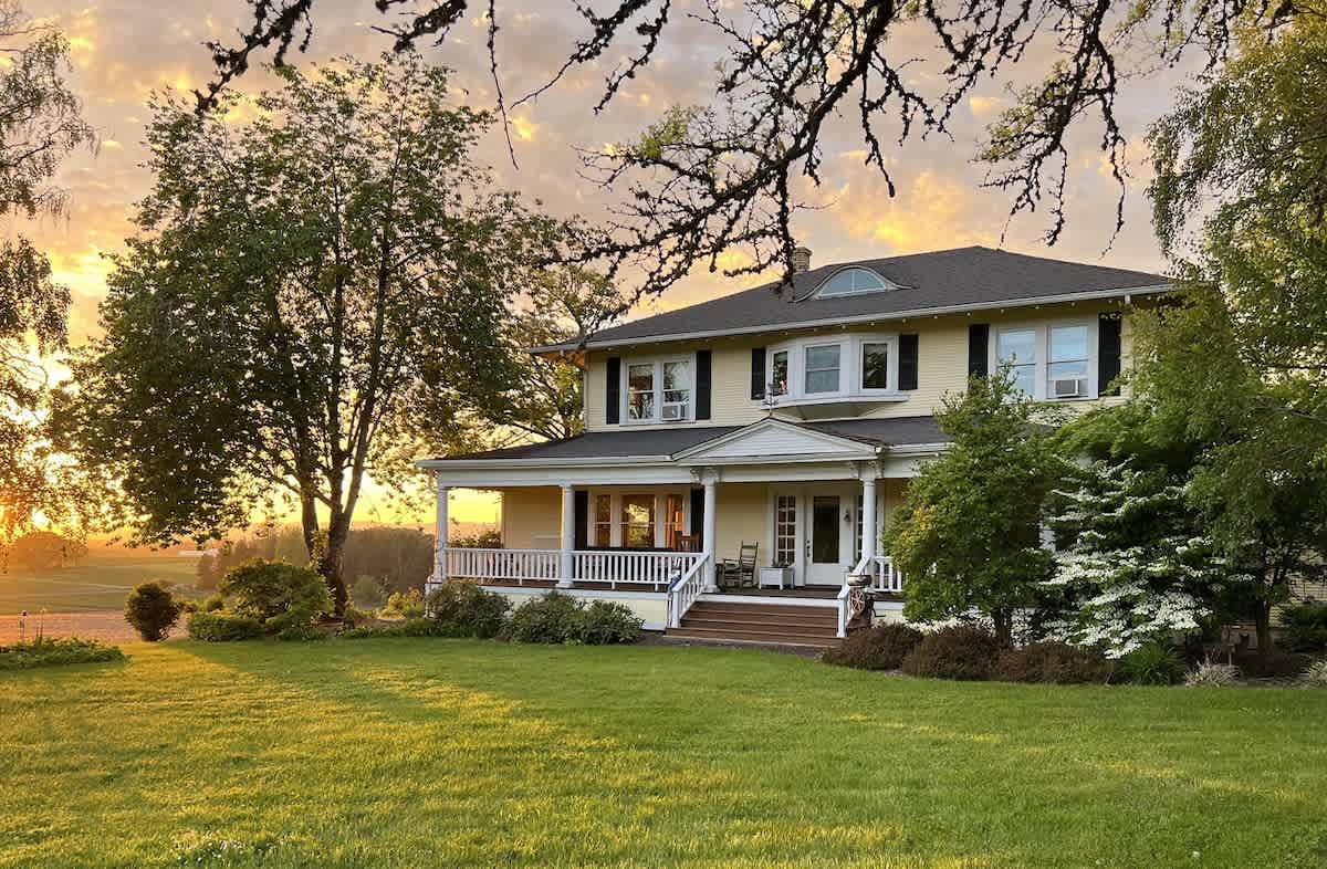 A grand yellow farm home with a dark roof and a wide front porch, surrounded by lush green lawn and mature trees, bathed in the warm glow of a sunset.
