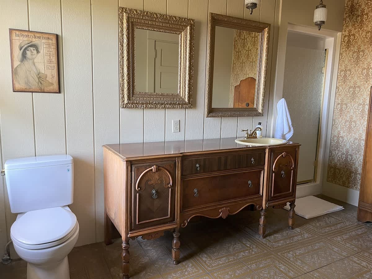 A vintage-style bathroom with a white toilet, a dark wood vanity with a cream sink, and two ornate gold-framed mirrors above it, set against paneled walls.