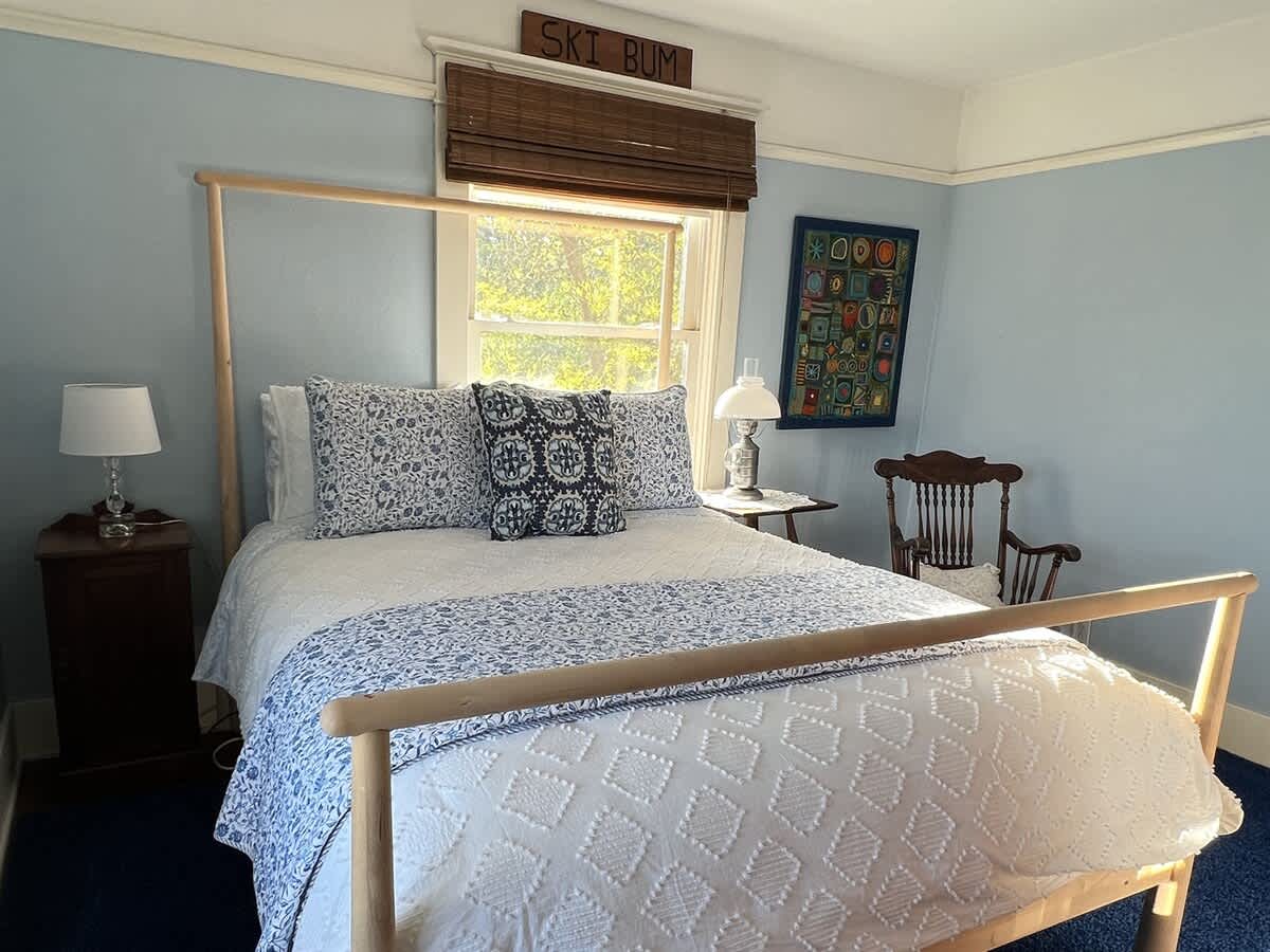 A close-up view of the light blue bedroom, highlighting the light wood bed frame with white and blue patterned bedding, a bedside table with a lamp, and a colorful abstract artwork on the wall. A "SKI BUM" sign is above the window.