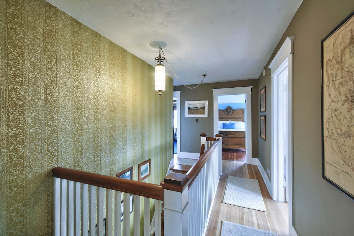 An upstairs hallway with a white banister and wooden handrail, featuring green patterned wallpaper and leading to a bedroom with a wooden bed frame.