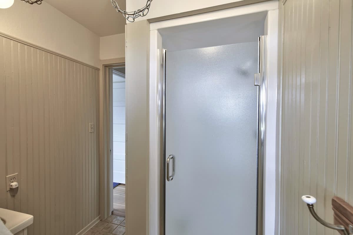 A bathroom featuring light-colored beadboard walls and a frosted glass shower stall with a chrome frame. A doorway leads to another part of the house, and a light fixture hangs from the ceiling.