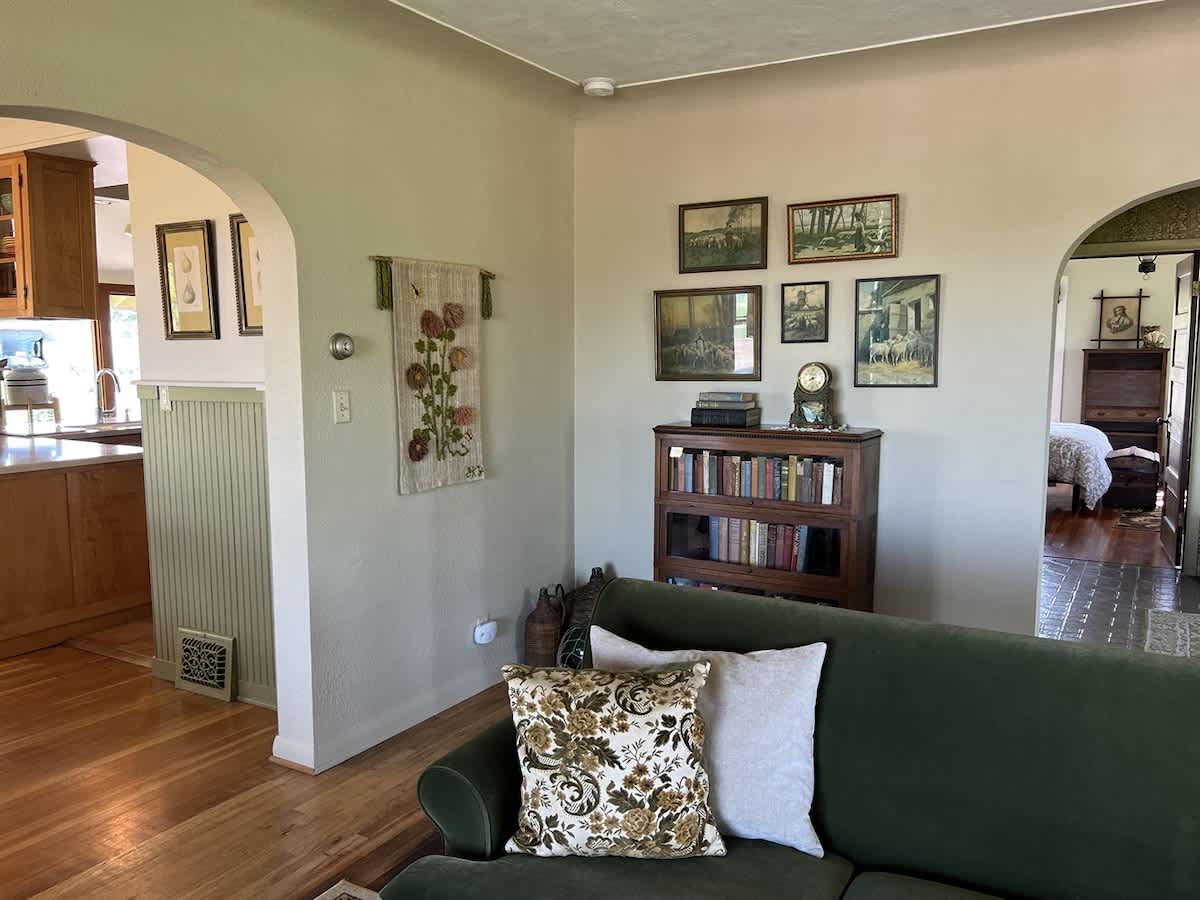 Traditional living room space with a green sofa adorned with decorative pillows, a wooden bookshelf filled with books, and a gallery wall of framed art, leading to an arched doorway with beadboard wainscoting.