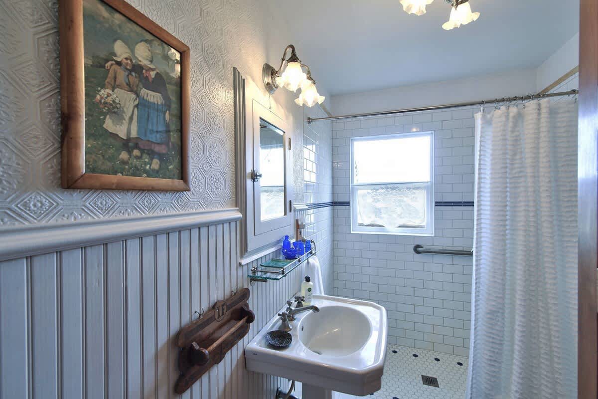 A bright bathroom with white subway tiles in the shower, a pedestal sink, and wainscoting with textured wallpaper above. The floor has small hexagonal tiles.