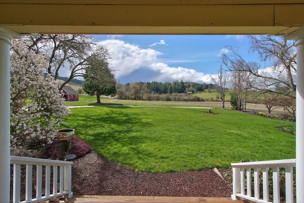 An expansive view from the porch reveals a vast green lawn leading to distant trees and a red barn, all under a bright blue sky adorned with large, fluffy white clouds.