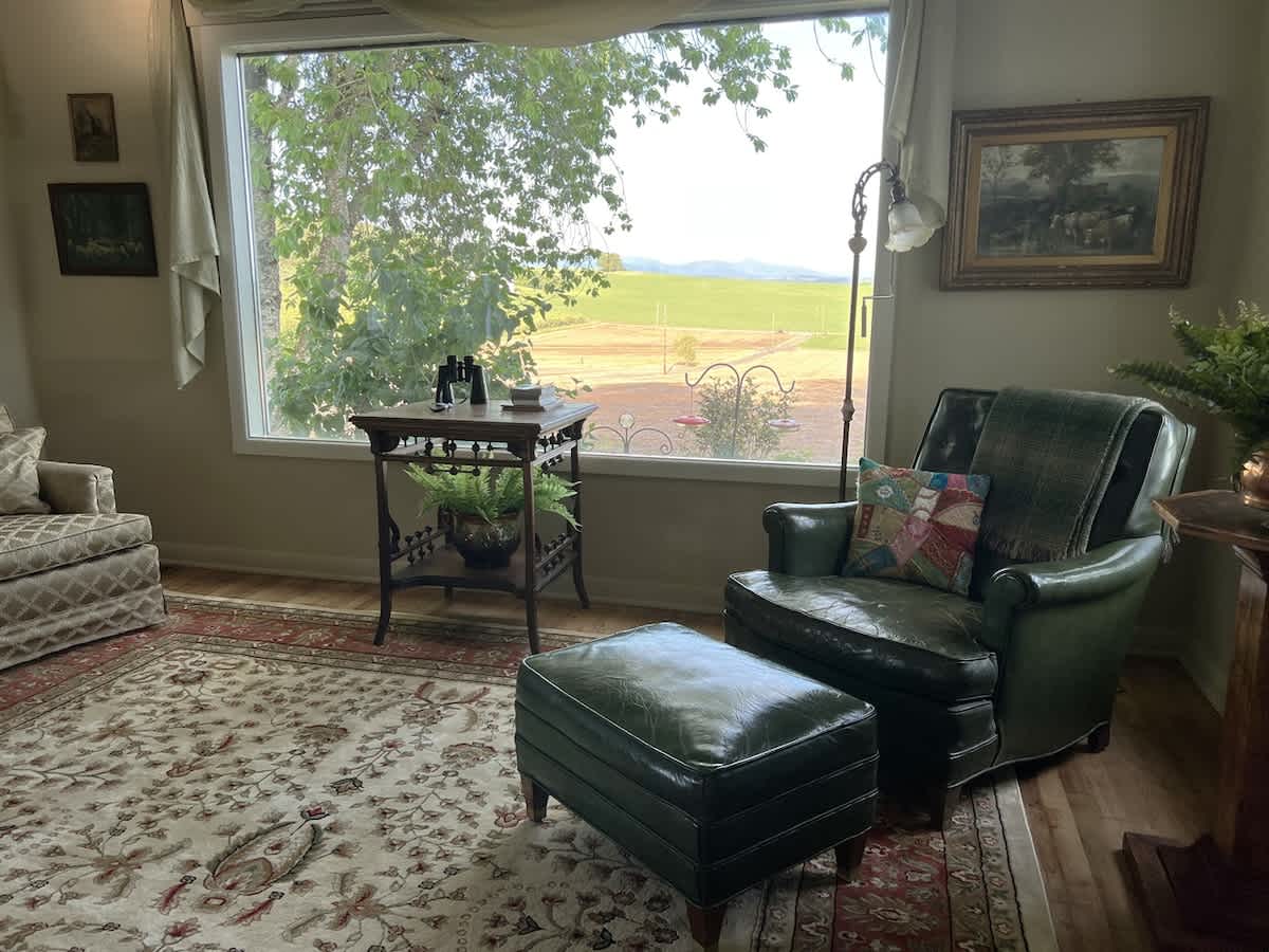 Inviting living room corner with a comfortable green armchair and matching ottoman, positioned by a large window that provides a scenic view of the surrounding fields and distant trees.