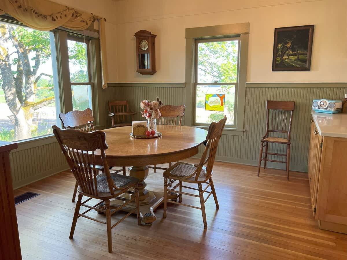 Traditional dining room with a round wooden table and chairs, featuring green beadboard wainscoting and light-colored walls, with a window providing natural light and a view of the outdoors.