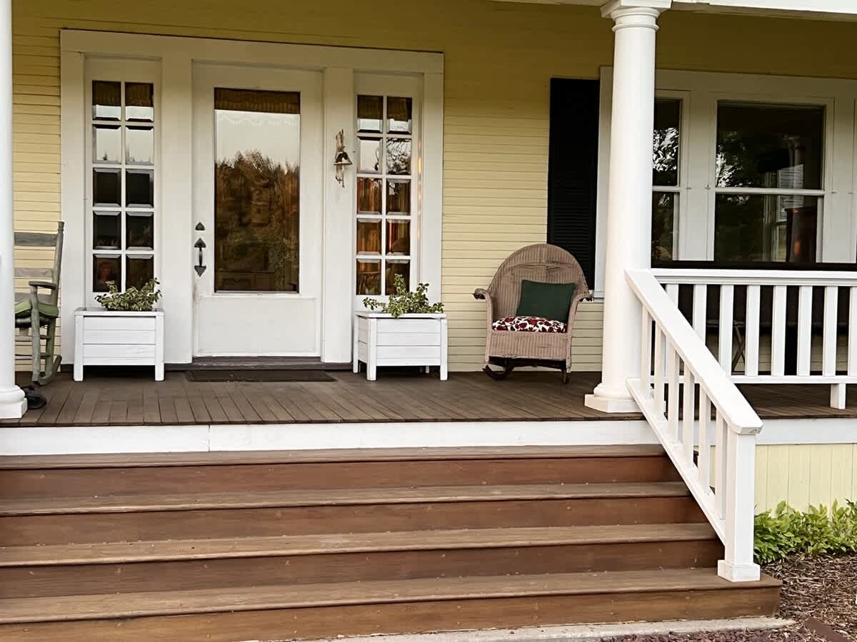 A welcoming front porch entrance features white double doors with glass panes, flanked by white planters, a comfortable wicker chair, and sturdy wooden steps leading up to the porch.