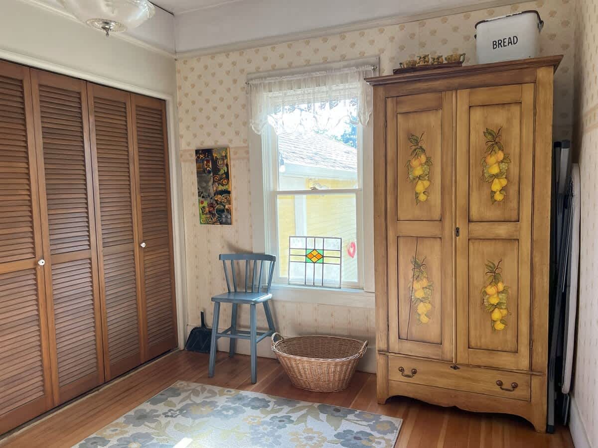 A room with light patterned wallpaper and hardwood floors, featuring a large painted wooden armoire with lemon motifs and louvered closet doors. A small window with a stained glass panel and a wicker basket are also visible.
