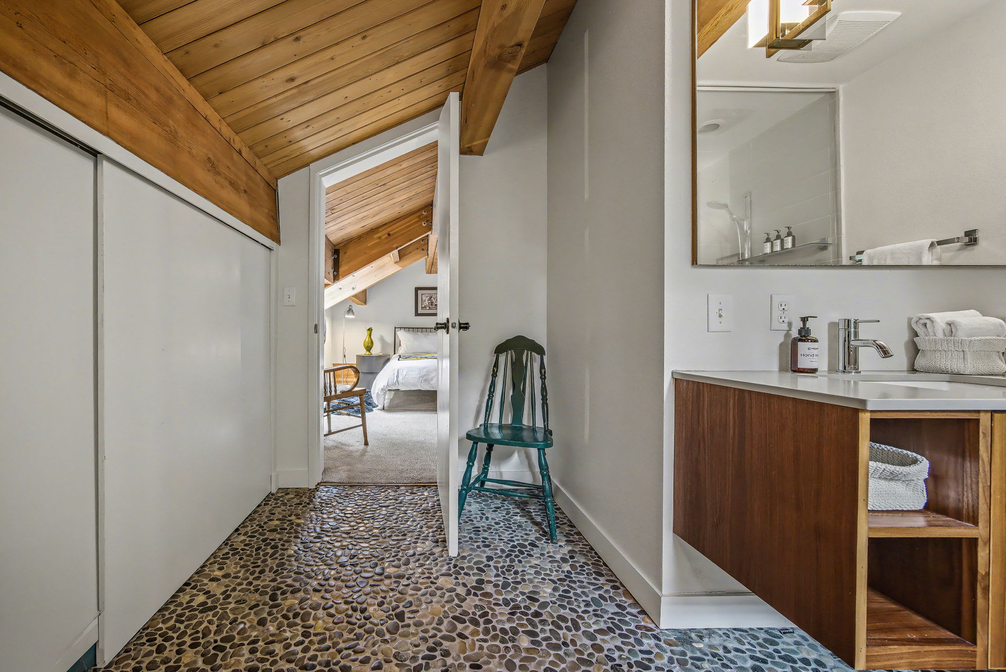 A hallway with a distinctive pebble-stone floor and a sloped wooden beam ceiling, leading to a bright bedroom with a bed and a bathroom with a modern vanity. A green wooden chair adds a pop of color to the space.