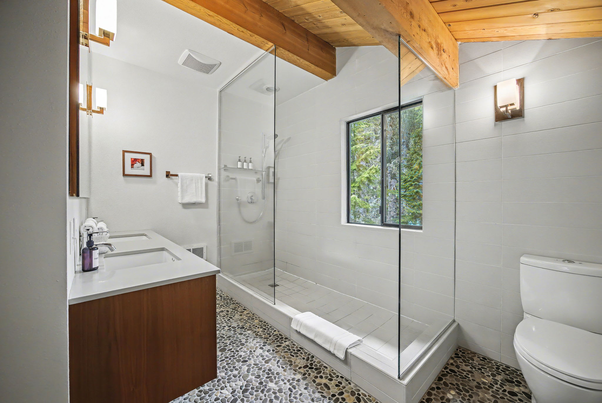 A bright bedroom with a white-linened bed and colorful blanket transitions into a bathroom through an open doorway, revealing dark pebble flooring. A wooden dresser with a mirror and a decorative chair are visible, all beneath a vaulted ceiling with exposed wooden beams.