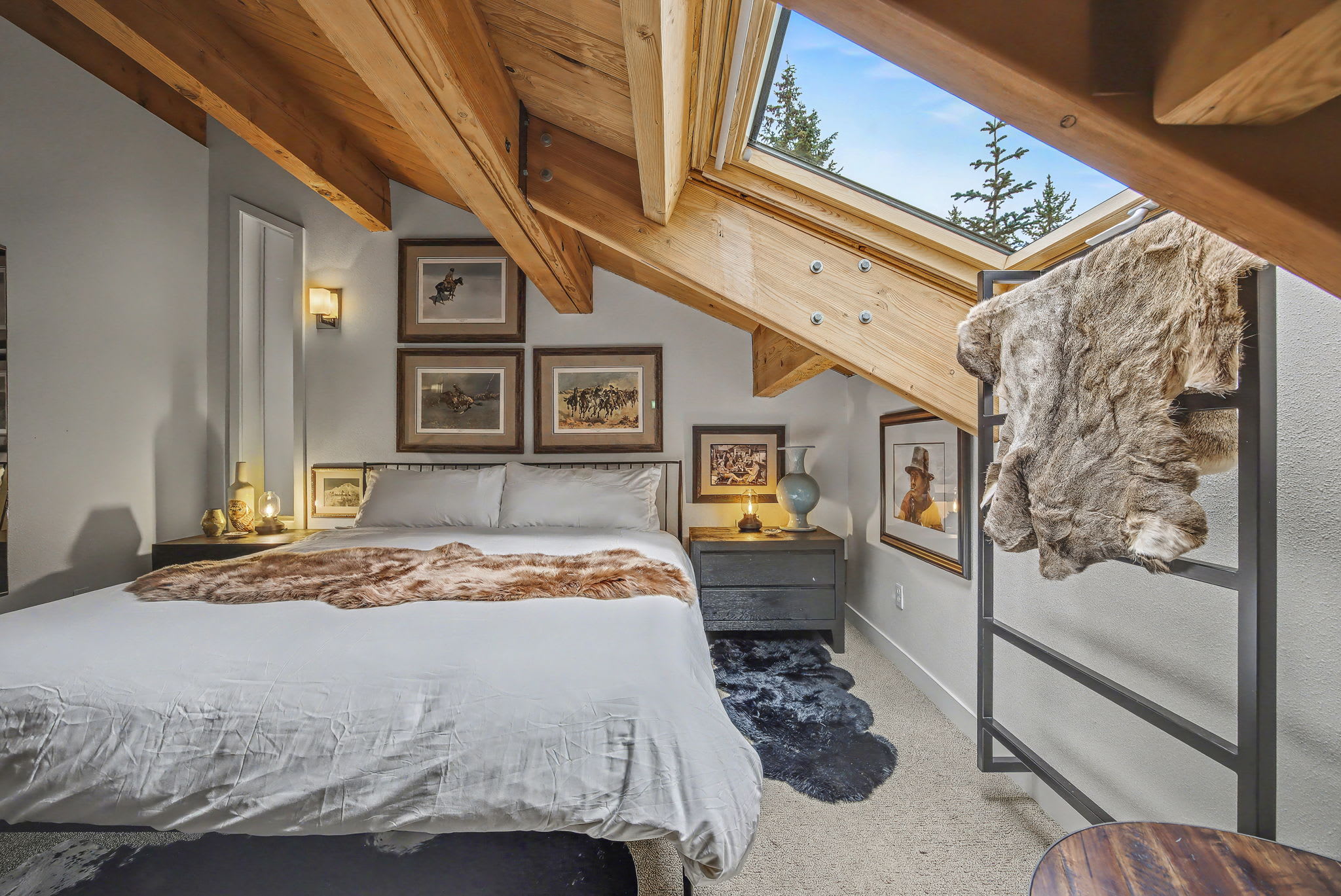 Spacious living room with warm hardwood floors, a large round leather ottoman, and a wood-paneled ceiling with exposed beams. Windows offer views of the surrounding evergreen trees.
