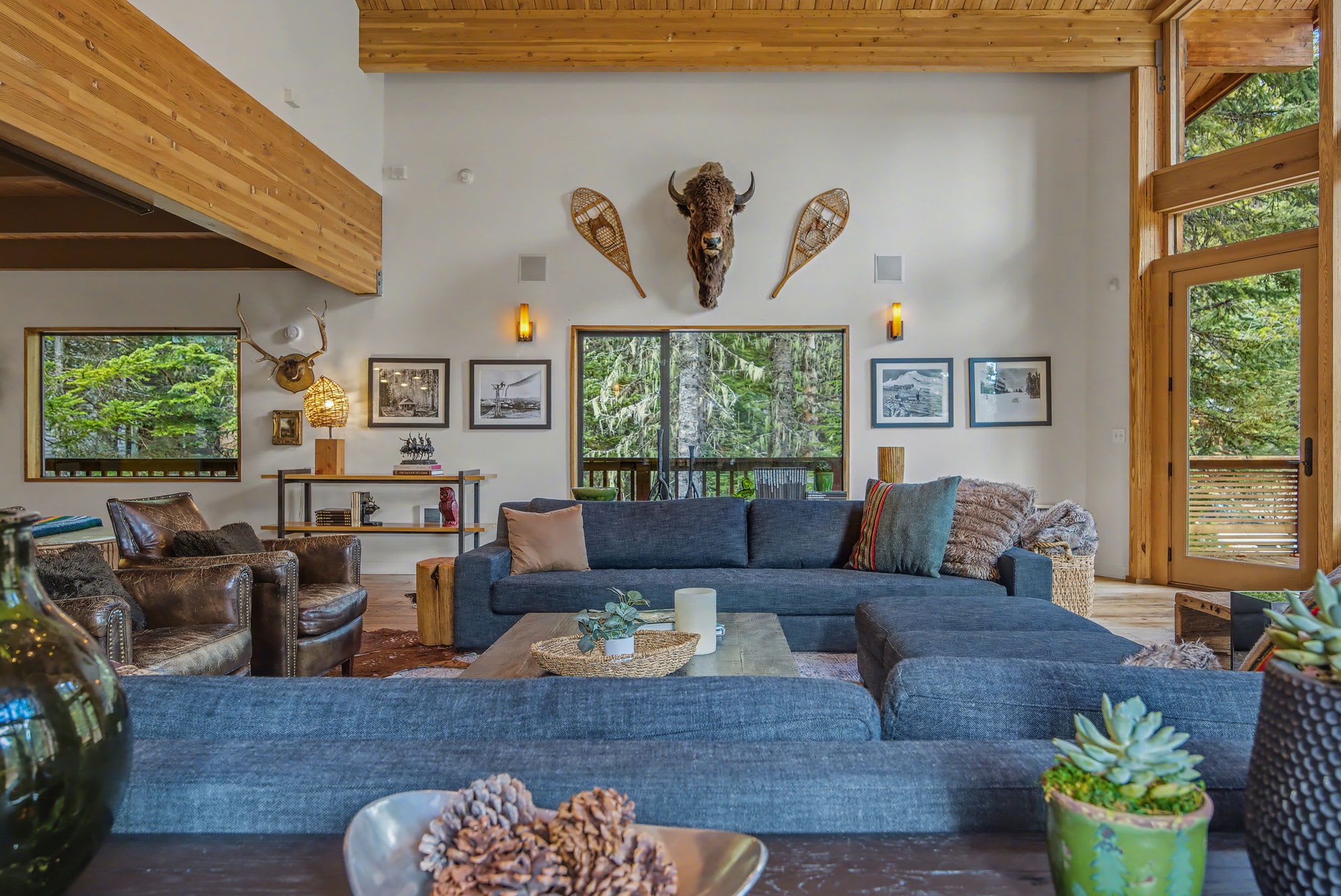 A grand living room with a soaring wood-paneled vaulted ceiling and exposed beams, centered around a massive stone fireplace. Multiple seating areas with dark sofas and leather armchairs are arranged on cowhide rugs, facing large windows with forest views.