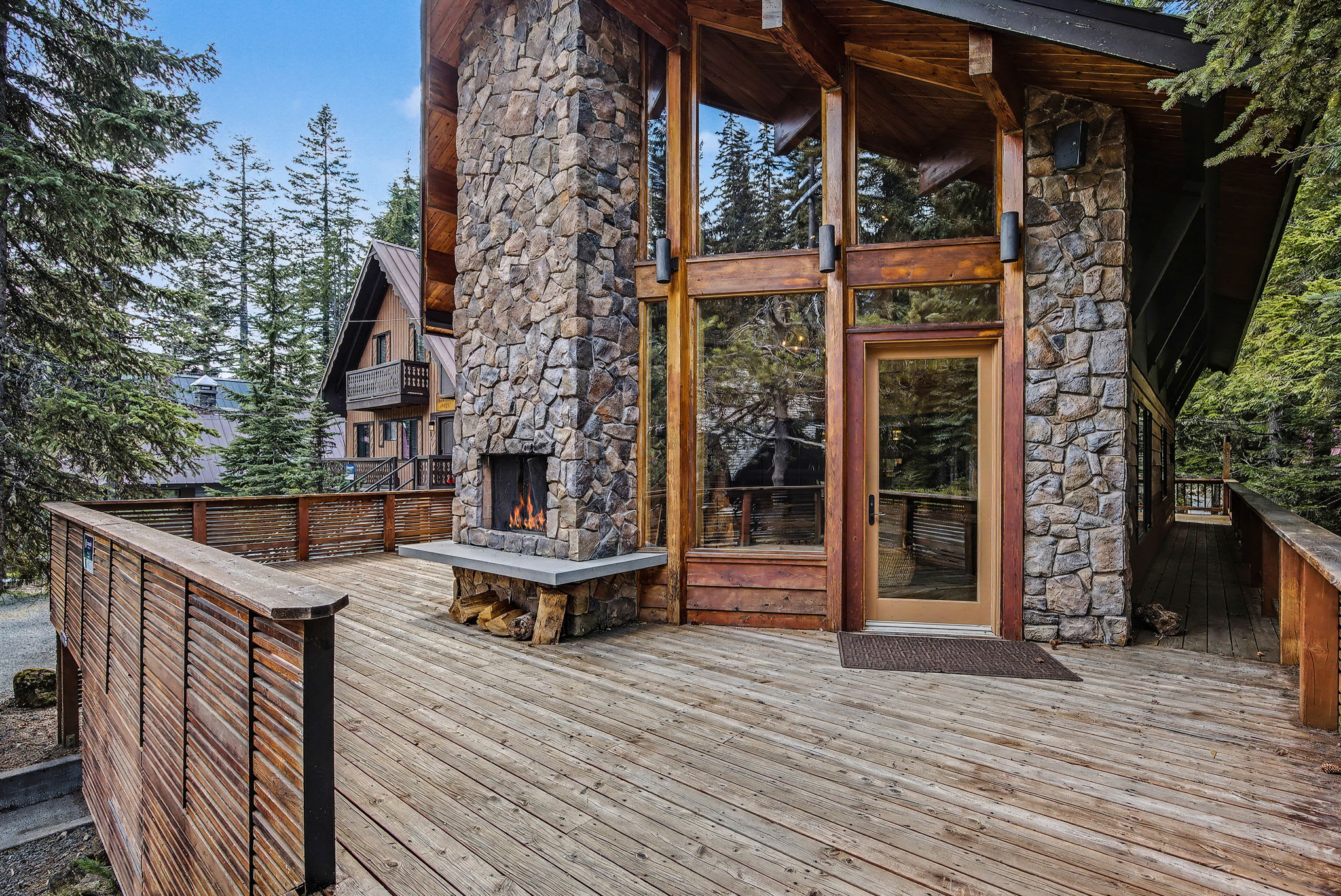 A spacious wooden deck wraps around the side of the chalet, featuring an outdoor fireplace built into the stone exterior. Large windows and a glass door connect the interior to the forest surroundings.